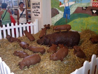 [Mama pig is made from dark chocolate and it surrounded by 13 milk chocolate piglets. All are set in hay and surrounded by a white picket fense with a painted farm scene background.]